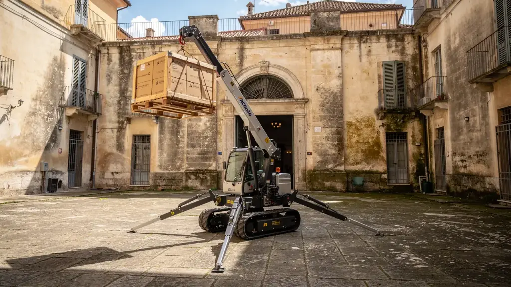 Gru ragno cingolata con stabilizzatori aperti che solleva una cassa nel cortile di un palazzo